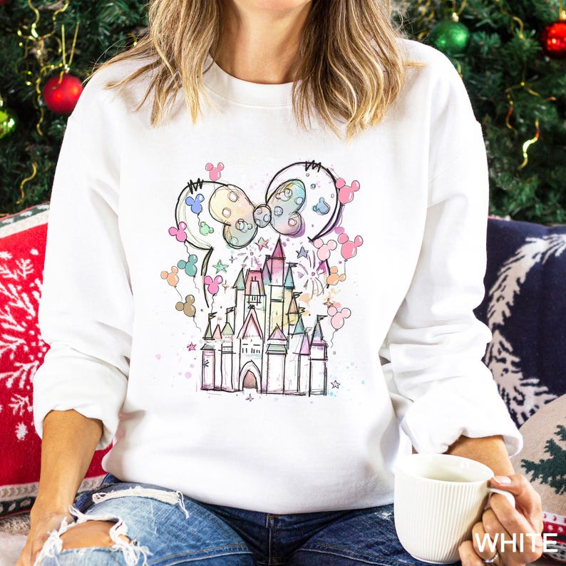 May include: White sweatshirt featuring a colorful castle design with a bow and Mickey Mouse ears. The graphic includes pastel colors and stars. The word "WHITE" is printed on the bottom right corner of the sweatshirt.
