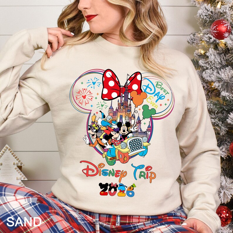 May include: A sand-colored sweatshirt featuring a colorful Disney-themed graphic. The graphic includes Mickey and Minnie Mouse, a castle, and the text "Disney Trip 2026". A red polka-dot bow and fireworks complete the design.