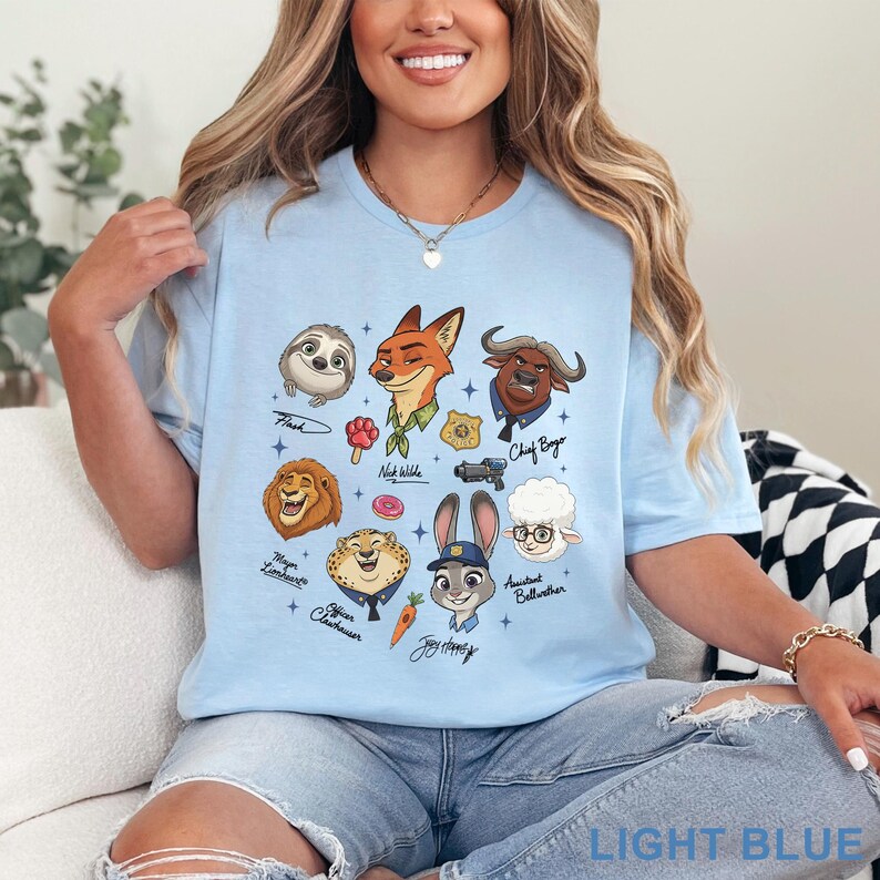 Pu&ograve; includere: T-shirt azzurra con personaggi dei cartoni animati di Zootropolis. Il design include Flash, Nick Wilde, Chief Bogo, Judy Hopps e altri personaggi. Il testo "LIGHT BLUE" &egrave; stampato in basso sulla maglietta.