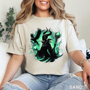 Puede incluir: Camiseta color arena con un gr&aacute;fico de una figura con cuernos, un cuervo y un bast&oacute;n. El dise&ntilde;o utiliza negro y tonos verdes. La palabra "SAND" est&aacute; impresa en la parte inferior de la camiseta.