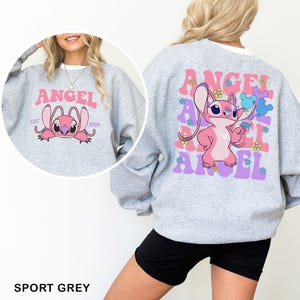 Puede incluir: Sudadera gris deportiva con un personaje de dibujos animados rosa y la palabra "ANGEL" en rosa y morado. La parte delantera de la sudadera tiene la cara del personaje y el texto "ANGEL EST 2004". La parte trasera tiene el personaje sosteniendo globos.