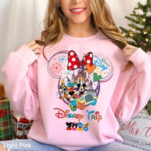 May include: Light pink sweatshirt featuring a colorful Disney Trip 2026 graphic. The design includes Mickey and Minnie Mouse, a castle, and the words "Best Day Ever." A large red bow with white polka dots is at the top of the design.