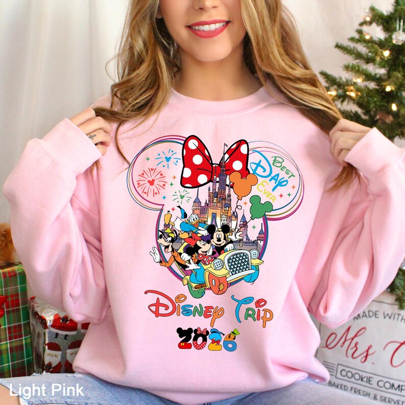 May include: Light pink sweatshirt featuring a colorful Disney Trip 2026 graphic. The design includes Mickey and Minnie Mouse, a castle, and the words "Best Day Ever." A large red bow with white polka dots is at the top of the design.