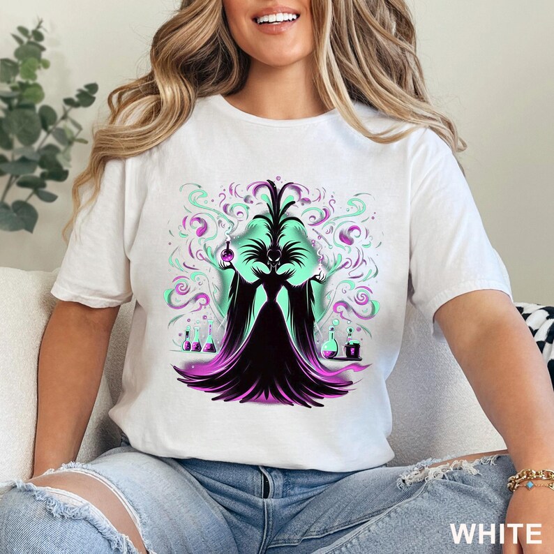 Puede incluir: Camiseta blanca con un gr&aacute;fico de una hechicera con un vestido negro, cara verde y acentos morados. El dise&ntilde;o incluye humo verde y morado arremolinado, botellas de pociones y la palabra "WHITE" en la parte inferior.