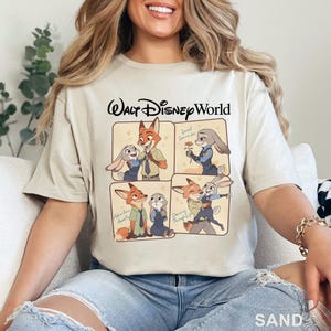 Puede incluir: Camiseta color arena con el logotipo de Walt Disney World y una ilustraci&oacute;n de cuatro paneles de personajes de Zootopia. Los paneles muestran a Nick Wilde y Judy Hopps en varias poses, con frases como "Sweet Surrender" y "Adventure Awaits".