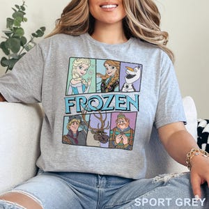 May include: A light gray t-shirt with a graphic print of characters from the movie Frozen. The characters include Elsa, Anna, Olaf, Kristoff, and Sven. The text "FROZEN" is printed in blue letters on the shirt. The shirt is available in the color Sport Grey.