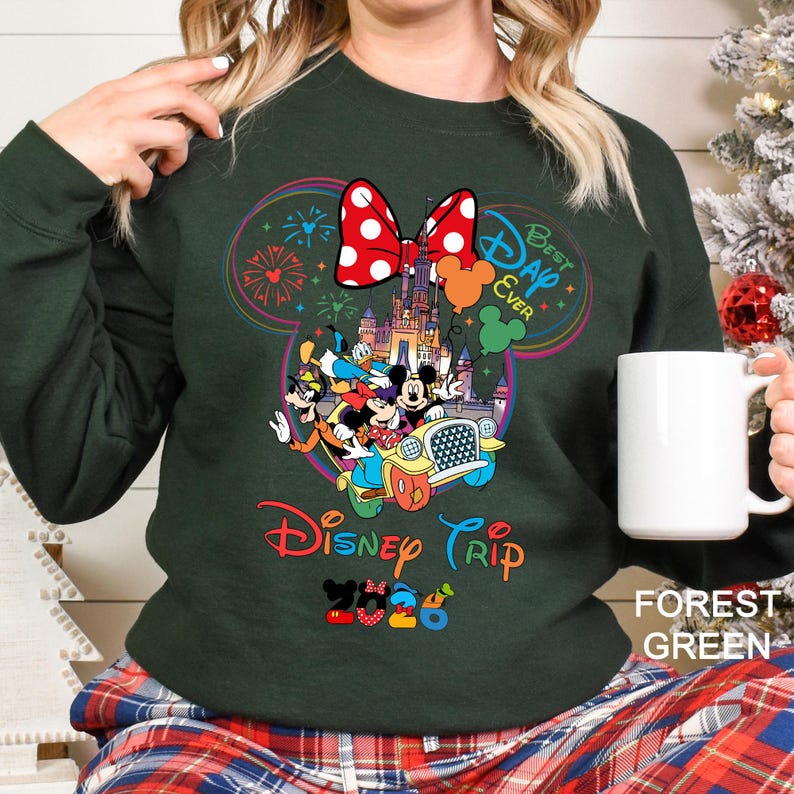 May include: Forest green sweatshirt with a colorful Disney Trip 2026 graphic. The design features Mickey and friends in a car, a castle, and the text "Best Day Ever". A large red bow with white polka dots is at the top.