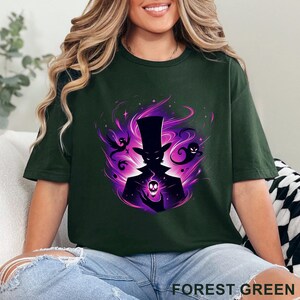 Puede incluir: Camiseta verde bosque con un gr&aacute;fico de una figura silueteada con sombrero de copa sosteniendo una calavera, rodeada de energ&iacute;a p&uacute;rpura y fantasmas. El texto "FOREST GREEN" est&aacute; impreso en la parte inferior.