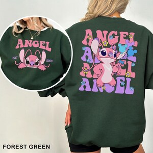 Puede incluir: Sudadera verde bosque con un dise&ntilde;o de personaje de dibujos animados rosa. La parte delantera presenta un gr&aacute;fico circular con la palabra "ANGEL" y el personaje. La parte trasera tiene un dise&ntilde;o "ANGEL" apilado con el personaje sosteniendo flores.