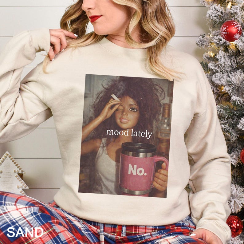 May include: A sand-coloured crewneck sweatshirt featuring a graphic print of a doll with messy hair holding a cigarette, with the text mood lately above a pink mug that has the word No. printed on it in white letters.