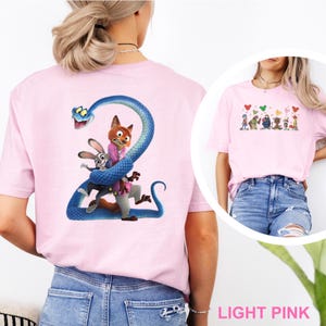 K&ouml;nnte beinhalten: Hellrosa T-Shirt mit einem Cartoon-Motiv eines Fuchses, Hasen und einer blauen Schlange. Der Fuchs tr&auml;gt eine rosa Jacke. Ein kleineres Bild von Cartoon-Charakteren ist ebenfalls vorhanden. Der Text "LIGHT PINK" befindet sich unten.