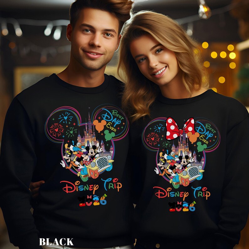 May include: Black sweatshirts featuring a colorful Disney Trip design. The design includes Mickey Mouse ears, fireworks, a castle, and the text "Best Day Ever." The sweatshirts are worn by a man and a woman.