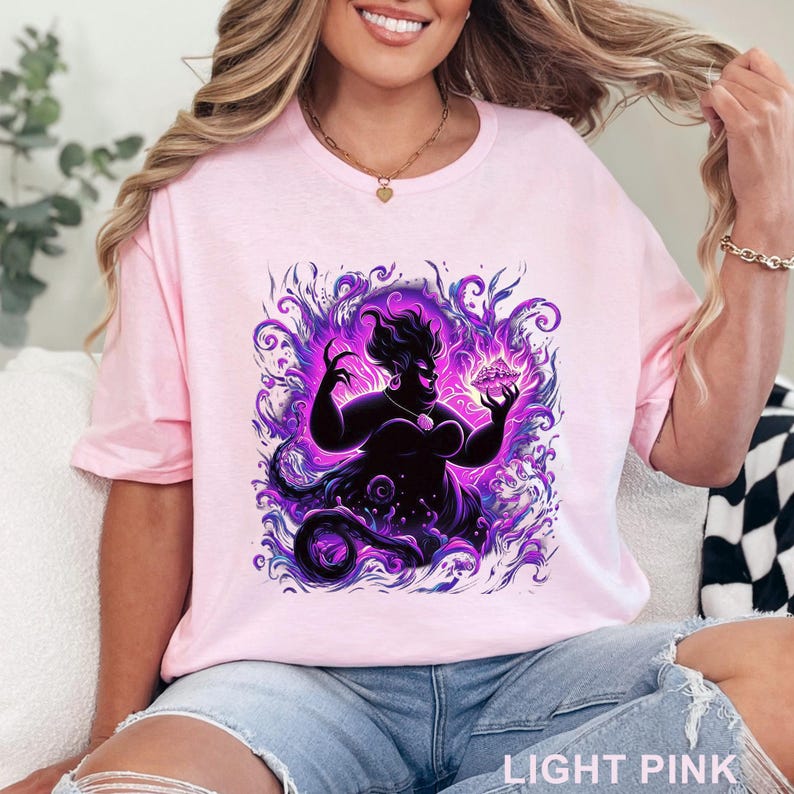 Puede incluir: Camiseta rosa claro con un gr&aacute;fico de un personaje oscuro con detalles morados y violetas. El personaje sostiene un objeto brillante. El texto "LIGHT PINK" est&aacute; en la parte inferior.