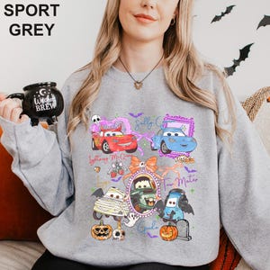 May include: A sport grey crewneck sweatshirt featuring a graphic print of Disney Pixar Cars characters in Halloween costumes, including Lightning McQueen, Sally Carrera, Mater, and Guido, surrounded by pumpkins, bats, and text labels.