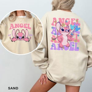 Puede incluir: Sudadera de color arena con un personaje de dibujos animados rosa y la palabra "ANGEL" en letras rosas y moradas. La parte delantera muestra la cara del personaje y la palabra "ANGEL". La parte trasera tiene el personaje sosteniendo globos. Estilo casual.