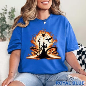 Puede incluir: Camiseta azul real con un gr&aacute;fico de una figura silueteada con un bast&oacute;n, sobre un fondo naranja y amarillo llameante con dos p&aacute;jaros negros. El texto "ROYAL BLUE" est&aacute; impreso en la parte inferior.