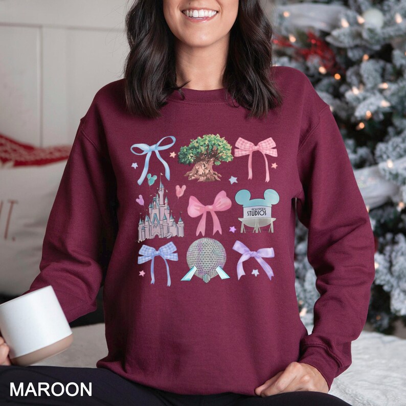 May include: Maroon sweatshirt featuring colorful illustrations of bows, a castle, a tree, and other theme park icons. The word "STUDIOS" is visible on one of the graphics. The word "MAROON" is printed at the bottom of the image.