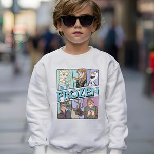 May include: White sweatshirt featuring a colorful graphic of characters from the movie "Frozen." The graphic includes Elsa, Anna, Olaf, Kristoff, Sven, and Hans. The word "FROZEN" is printed in blue letters below the characters.
