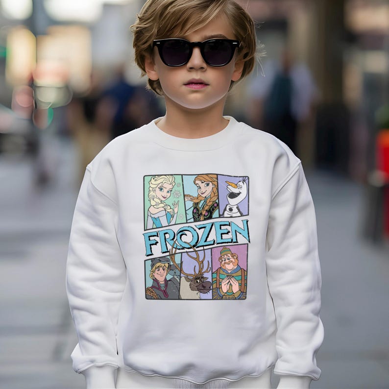 May include: White sweatshirt featuring a colorful graphic of characters from the movie "Frozen." The graphic includes Elsa, Anna, Olaf, Kristoff, Sven, and Hans. The word "FROZEN" is printed in blue letters below the characters.