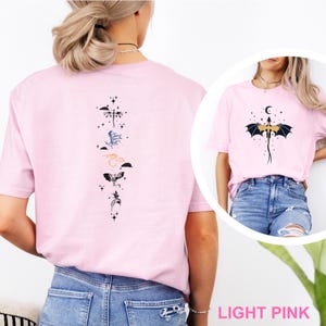 Pu&ograve; includere: T-shirt rosa chiaro con disegni di draghi sul davanti e sul retro. Il retro presenta una disposizione verticale di draghi stilizzati in vari colori. Il design frontale mostra un drago con una luna crescente e stelle. Il testo "LIGHT PINK" &egrave; in basso.