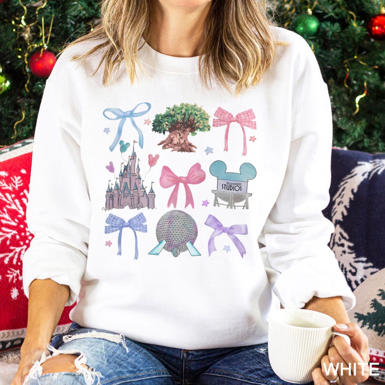 May include: White sweatshirt featuring colorful illustrations of a castle, tree, bows, and other theme park icons. The design includes the text "STUDIOS" in a blue Mickey Mouse head. The word "WHITE" is printed on the bottom right.