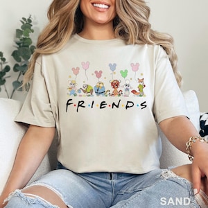 May include: Sand-colored t-shirt featuring a cartoon design of various characters holding Mickey Mouse-shaped balloons. The word "FRIENDS" is printed in black below the characters. The shirt is made of a soft material.