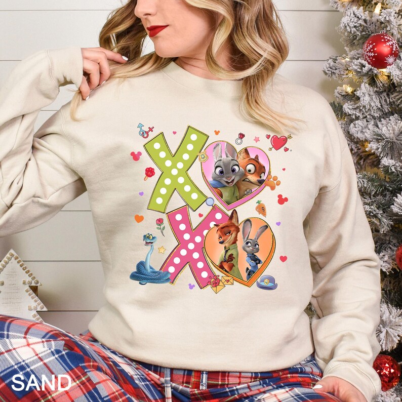 May include: A sand-colored sweatshirt featuring a graphic design with the letters "XO" in green and pink, decorated with cartoon characters in heart-shaped frames. The design includes cartoon animals and other decorative elements.