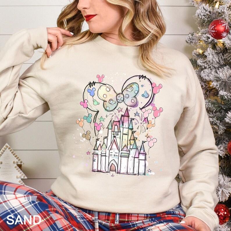 May include: Sand-colored sweatshirt featuring a colorful castle design with a bow and mouse ears. The graphic includes pastel colors and stars. The sweatshirt is long-sleeved and has a crew neck.