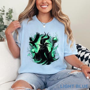Puede incluir: Camiseta azul claro con un gr&aacute;fico de una silueta con cuernos, un cuervo y un bast&oacute;n con un orbe verde. El dise&ntilde;o est&aacute; rodeado de remolinos verdes. El texto "LIGHT BLUE" est&aacute; en la parte inferior de la camiseta.