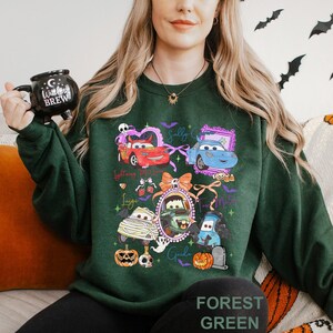 May include: A forest green crewneck sweatshirt featuring a colourful graphic print of various animated car characters in Halloween costumes, including pumpkins, bats, and ghosts, with names like Lightning McQueen, Sally, and Mater printed.