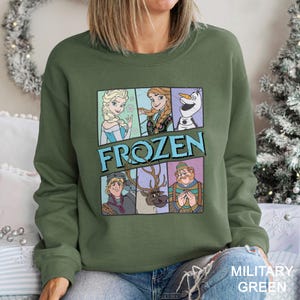 May include: Military green sweatshirt featuring a graphic of characters from the movie "Frozen." The design includes Elsa, Anna, Olaf, Kristoff, Sven, and Hans. The word "FROZEN" is displayed in large, blue letters. The sweatshirt is a crew neck style.