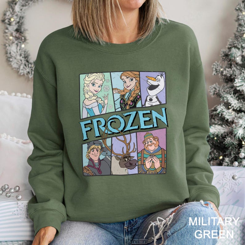May include: Military green sweatshirt featuring a graphic of characters from the movie "Frozen." The design includes Elsa, Anna, Olaf, Kristoff, Sven, and Hans. The word "FROZEN" is displayed in large, blue letters. The sweatshirt is a crew neck style.