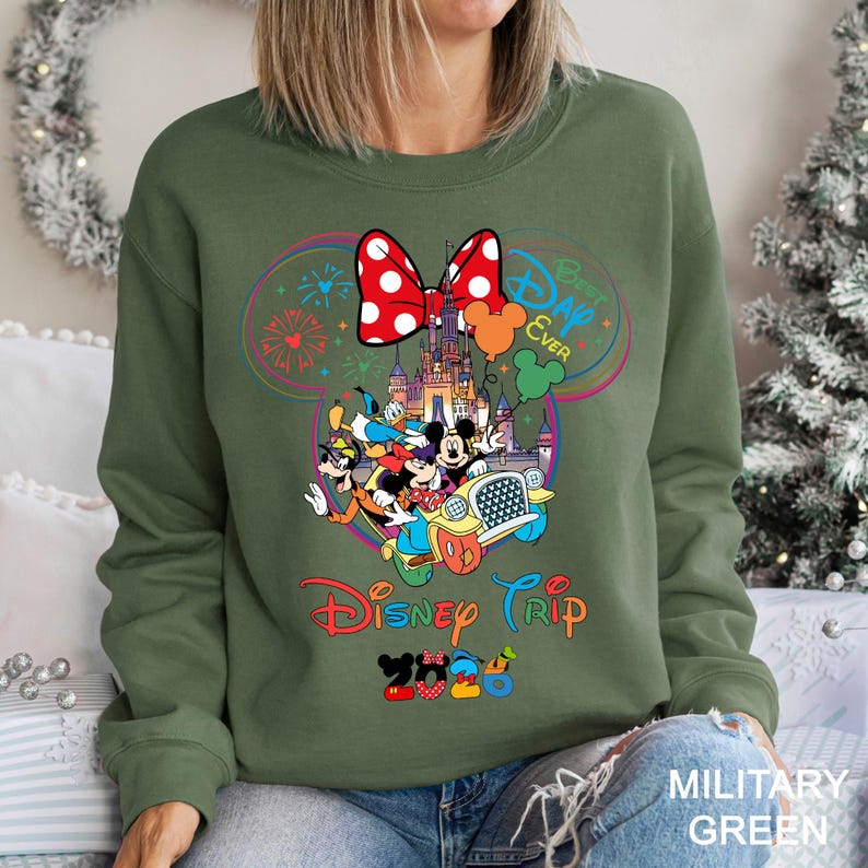 May include: Military green sweatshirt featuring a colorful Disney graphic. The design includes Mickey Mouse, Donald Duck, and other characters in a car, with text that reads "Disney Trip 2023" and "Best Day Ever". A large red bow with white polka dots is at the top.