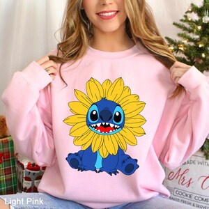 May include: A light pink long-sleeve sweatshirt featuring a graphic print of the blue character Stitch from Lilo and Stitch, depicted with a large yellow sunflower surrounding its head, showcasing a wide grin with visible white teeth.