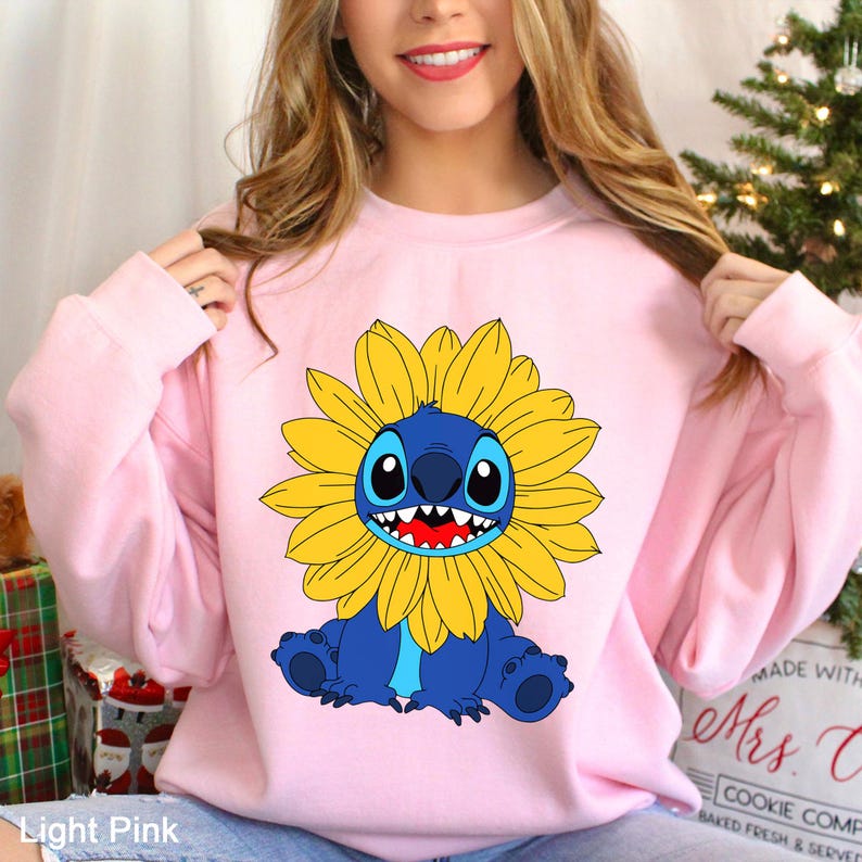 May include: A light pink long-sleeve sweatshirt featuring a graphic print of the blue character Stitch from Lilo and Stitch, depicted with a large yellow sunflower surrounding its head, showcasing a wide grin with visible white teeth.