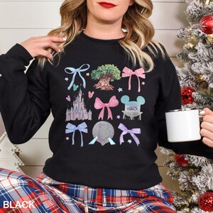 May include: Black crewneck sweatshirt featuring colorful illustrations of bows, a castle, a tree, and other theme park icons. The word "STUDIOS" is visible on the design. The sweatshirt is worn by a person.