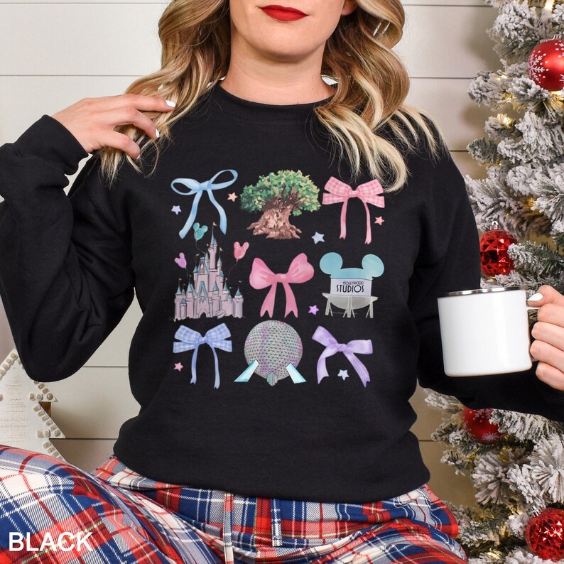 May include: Black crewneck sweatshirt featuring colorful illustrations of bows, a castle, a tree, and other theme park icons. The word "STUDIOS" is visible on the design. The sweatshirt is worn by a person.