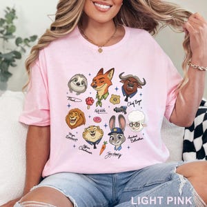 Pu&ograve; includere: T-shirt rosa chiaro con personaggi dei cartoni animati di Zootropolis. Il design include Flash, Nick Wilde, Chief Bogo, Mayor Lionheart, Officer Clawhauser, Judy Hopps e altri. Il testo "LIGHT PINK" &egrave; in basso.