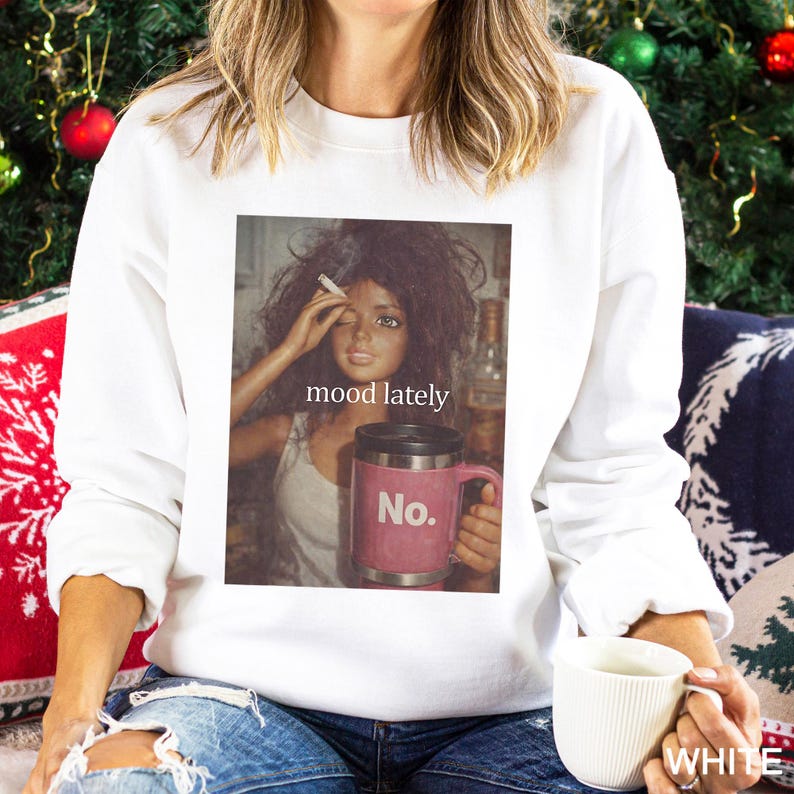 May include: A white long-sleeve sweatshirt featuring a graphic print of a doll with messy hair holding a cigarette and a pink travel mug with the text No. The text mood lately is printed above the mug. The sweatshirt has a crew neckline.