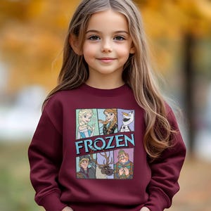 May include: A young girl wearing a maroon sweatshirt featuring a colorful graphic from the movie "Frozen." The graphic includes images of Elsa, Anna, Olaf, Kristoff, and Sven, with the word "FROZEN" in blue letters.
