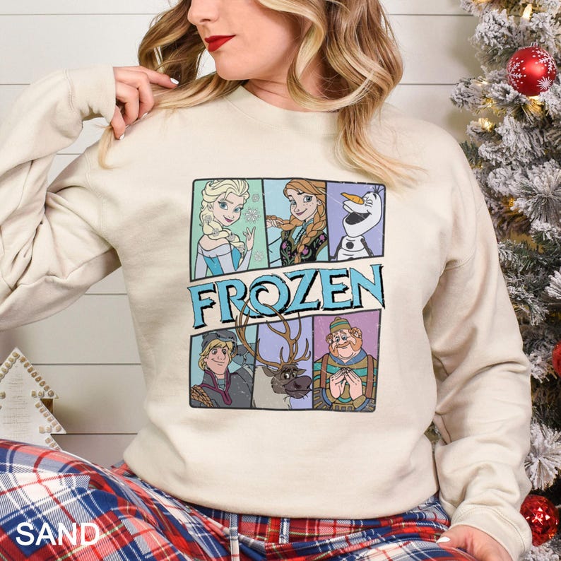 May include: A sand-colored sweatshirt featuring a colorful graphic of characters from the movie "Frozen." The graphic includes Elsa, Anna, Olaf, Kristoff, Sven, and a character in a blue and green striped sweater. The word "FROZEN" is printed in blue.