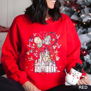 May include: A red sweatshirt featuring a castle design with a bow and mouse ears. The castle is in pastel colors, and the bow has a polka dot pattern. The design is surrounded by colorful splatters and mouse silhouettes.