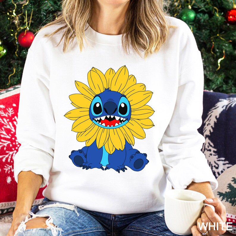 May include: A white long-sleeve sweatshirt featuring a graphic print of a blue creature with large black eyes and a wide grin, surrounded by a bright yellow sunflower-shaped mane. The design is centered on the chest of the garment.