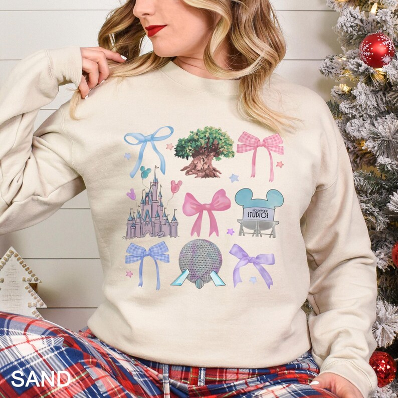 May include: Sand-colored sweatshirt featuring a colorful graphic design of bows, a castle, a tree, and other theme park icons. The word "STUDIOS" is visible in the design. The sweatshirt has long sleeves.