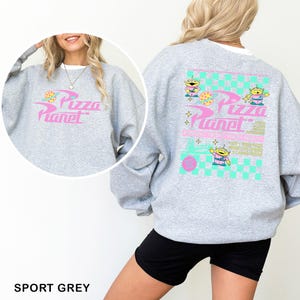 May include: Sport grey sweatshirt featuring the Pizza Planet logo in pink and blue on the front and back. The back design includes cartoon aliens and a checkered pattern. The sweatshirt is a casual, comfortable style.