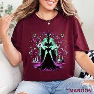 Puede incluir: Camiseta granate con un gr&aacute;fico de una figura estilizada con un vestido negro y una cara verde, rodeada de humo morado y verde arremolinado y frascos de laboratorio. La palabra "MAROON" est&aacute; impresa en la parte inferior.
