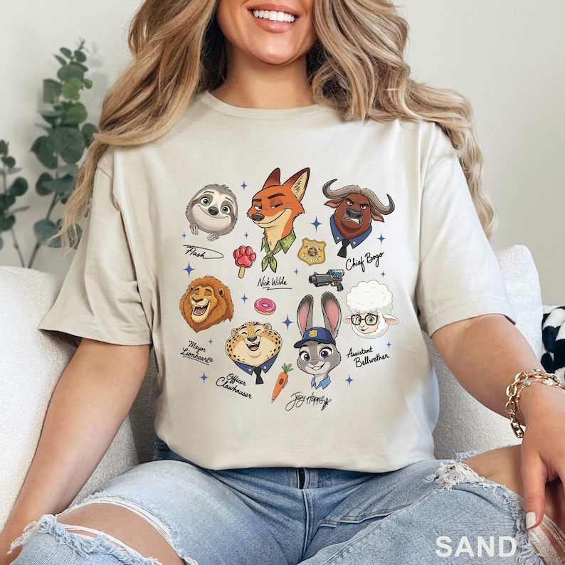Pu&ograve; includere: T-shirt color sabbia con illustrazioni di personaggi del film Zootropolis. Il design include Flash il bradipo, Nick Wilde la volpe, il capo Bogo il bufalo e altri. La parola "SAND" &egrave; stampata in basso.