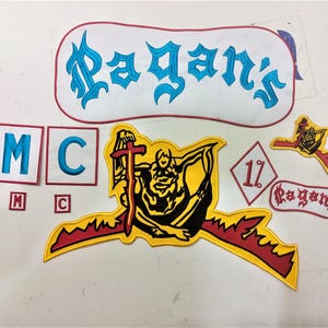 Może przedstawiać: Kolekcja haftowanych naszywek z różnymi wzorami i napisami. Naszywki zawierają napis "Pagan's" w niebieskim skrypcie, "MC" w kolorze niebieskim oraz naszywkę w kształcie rombu z napisem "1%" w kolorze czerwonym. Inne naszywki przedstawiają postać z krzyżem.