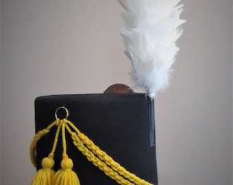 May include: A black hat with a flat brim, adorned with a large white feather plume. The hat features a yellow braided cord and two yellow tassels. A gold ring is attached to the hat.