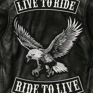 May include: Black leather jacket with a large embroidered eagle patch. The patch features a detailed white and black eagle with outstretched wings. Above the eagle is a white banner that reads "LIVE TO RIDE", and below it, another banner that reads "RIDE TO LIVE".